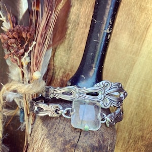 Peut inclure: Deux bracelets en argent fabriqués à partir d'argenterie recyclée, dont l'un avec une pierre précieuse carrée et transparente. Les bracelets sont présentés sur une surface en bois avec une poignée sombre et texturée en arrière-plan. Un bouquet de plumes et de fleurs séchées est également visible.