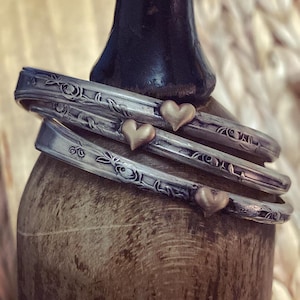 Puede incluir: Tres pulseras de plata con diseños florales y detalles en forma de corazón de oro. Las pulseras están apiladas en un expositor de madera.
