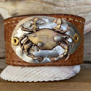 May include: A brown leather cuff bracelet with a silver and gold metal crab design. The crab is raised and has a textured surface.