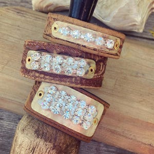 Puede incluir: Tres pulseras de cuero apiladas con textura de piel de serpiente. Cada pulsera tiene una placa rectangular de latón adornada con múltiples pedrerías claras y brillantes. El cuero es de color marrón cálido y las placas de latón están aseguradas con pequeños remaches dorados.