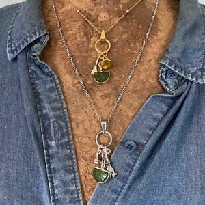 May include: Two layered necklaces featuring delicate chains. The shorter gold-tone chain holds a circular ring with small charms and a green slice-shaped pendant. The longer silver-tone chain features a matching ring with charms and a green pendant.