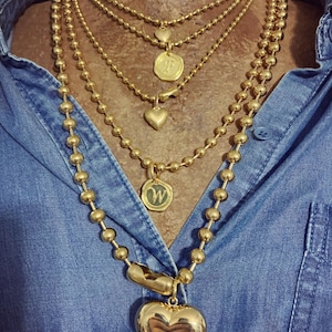 May include: A selection of gold-coloured necklaces with different pendant designs. The necklaces feature ball chains and heart-shaped pendants, along with other charms. The necklaces are layered and worn over a blue denim shirt.