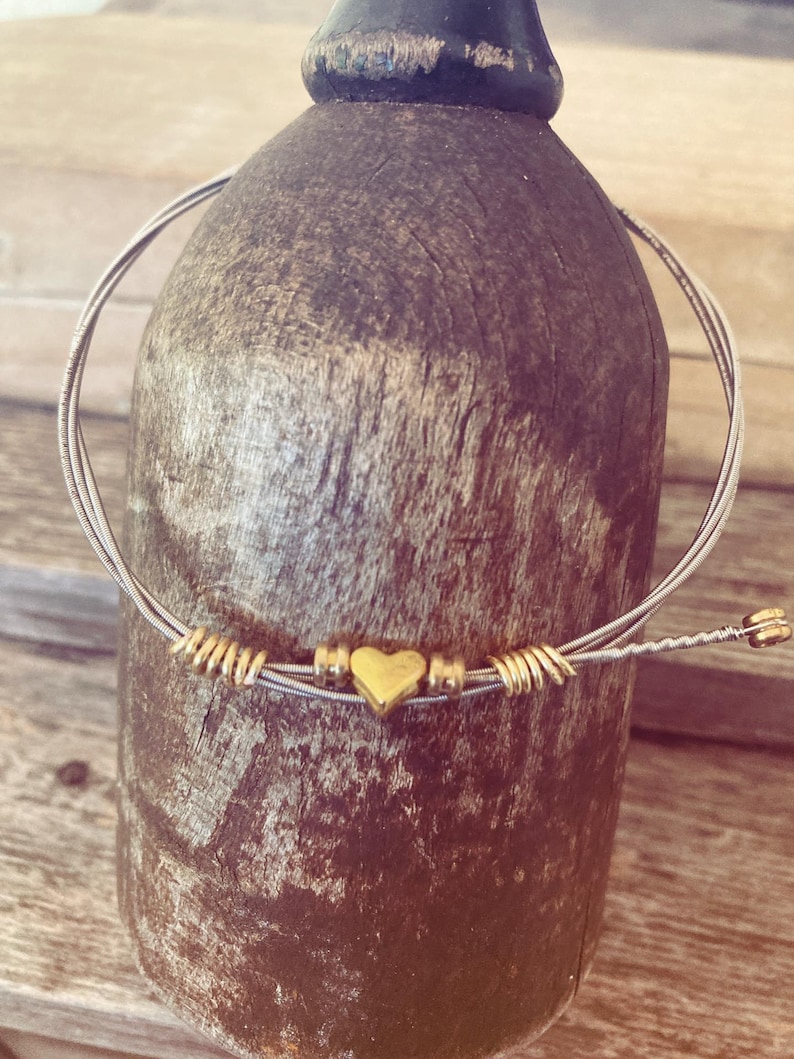 May include: A handmade bracelet featuring a silver-colored string with gold-colored accents. The bracelet has a small gold heart charm in the center and is wrapped around a wooden object. The bracelet is a unique piece of jewelry.