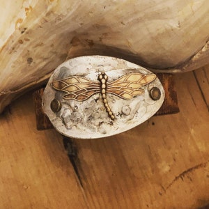 Op de afbeelding: Een handgemaakte metalen armband met een libellenontwerp. De ovale, zilverkleurige metalen plaat heeft een getextureerd oppervlak en is versierd met een goudkleurige libel. De armband is bevestigd aan een bruine leren band en vastgezet met messing klinknagels.
