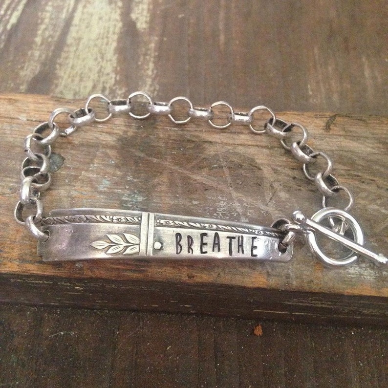 May include: Silver bracelet with a chain and a tag that says "BREATHE" with a leaf design.