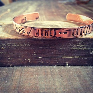 May include: A copper bracelet engraved with the words "Loved Women Rarely". The bracelet features a small arrow pointing to the right.