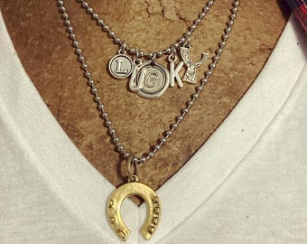 May include: A silver chain necklace with multiple charms, including a horseshoe, a letter 'L', a letter 'C', a letter 'K', and a letter 'Y'.