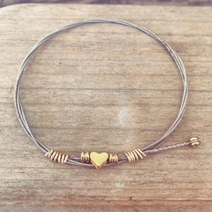 May include: A handmade bracelet crafted from silver-colored guitar strings, featuring gold-colored accents and a heart-shaped charm. The bracelet is displayed on a wooden surface, showcasing its unique design and craftsmanship.