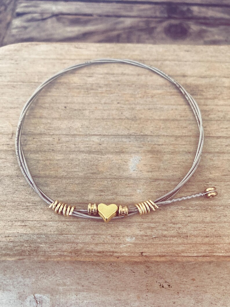 May include: A handmade bracelet crafted from silver-colored guitar strings, featuring gold-colored accents and a heart-shaped charm. The bracelet is displayed on a wooden surface, showcasing its unique design and craftsmanship.