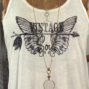 May include: A white tank top with a black graphic of wings and an arrow with the text "Vintage Soul". The model is wearing two necklaces, one with a silver locket and the other with a gold magnifying glass pendant.