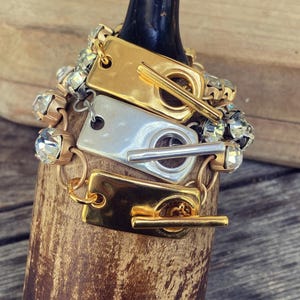 May include: A stack of bracelets with gold and silver rectangular clasps and clear crystal accents. The bracelets are draped over a wooden object with a dark handle. The bracelets have a toggle clasp closure.