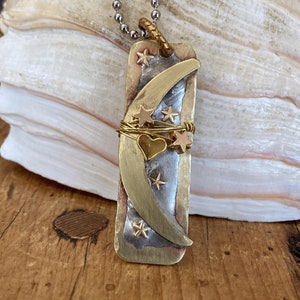 May include: A silver chain necklace with a brass pendant featuring a crescent moon, a heart, and stars. The pendant is made of mixed metals and has a rustic, handmade look.
