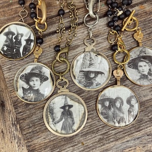 May include: A collection of vintage-style necklaces featuring round pendants with black and white images of women in witch hats. The pendants are set in gold-toned metal frames and hang from various chains with black bead accents.