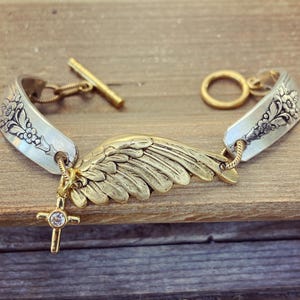 May include: A silver spoon bracelet with floral detailing and a gold-colored wing charm. The bracelet features a gold cross pendant with a clear stone and a toggle clasp. The bracelet is displayed on a wooden surface.