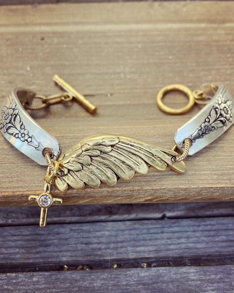 May include: A silver spoon bracelet with floral detailing and a gold-colored wing charm. The bracelet features a gold cross pendant with a clear stone and a toggle clasp. The bracelet is displayed on a wooden surface.
