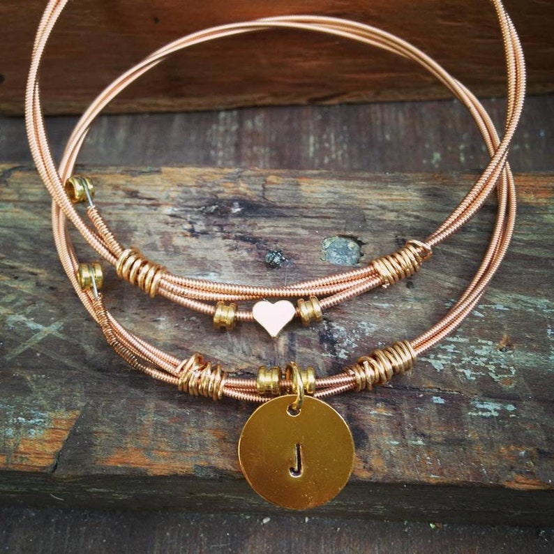 May include: A layered copper-toned necklace with a heart-shaped charm and a circular pendant. The pendant features the letter "J". The necklace is made of coiled wire and has a rustic, handmade appearance.