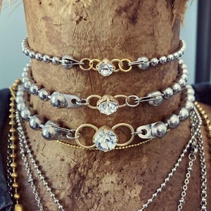 May include: A collection of necklaces and chokers. The chokers feature silver ball chains with gold-toned accents and a central clear crystal. Additional necklaces include silver and gold-toned chains of varying styles, creating a layered look.