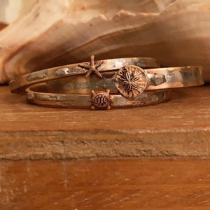 Può includere: Tre braccialetti in argento e rame con una stella marina, un dollaro di sabbia e un ciondolo a forma di tartaruga. I braccialetti sono impilati uno sopra l'altro e sono su una superficie di legno.