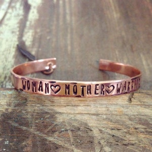 May include: Copper bracelet with the words "WOMAN MOTHER WARRIOR" engraved on it, with a heart symbol between each word.