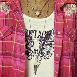 May include: A white t-shirt with the text "Vintage Soul" and a graphic of wings. The shirt is layered under a red and black plaid flannel shirt with lace trim. The person is wearing two necklaces, one with a heart pendant and the other with a wing pendant.