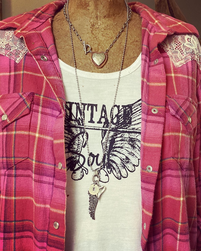 May include: A white t-shirt with the text "Vintage Soul" and a graphic of wings. The shirt is layered under a red and black plaid flannel shirt with lace trim. The person is wearing two necklaces, one with a heart pendant and the other with a wing pendant.
