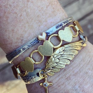 May include: A stack of gold and silver bracelets. One bracelet has heart-shaped links, another has a wing charm, and a third has a cross pendant with a small gem. The bracelets are worn together.
