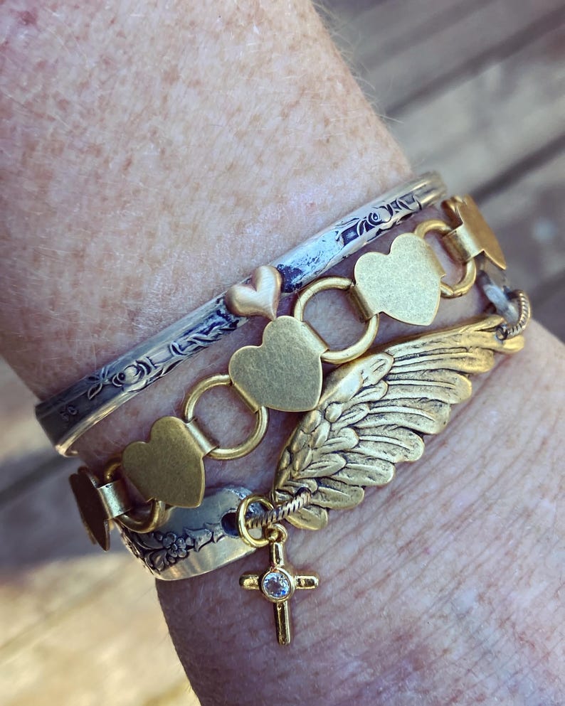 May include: A stack of gold and silver bracelets. One bracelet has heart-shaped links, another has a wing charm, and a third has a cross pendant with a small gem. The bracelets are worn together.