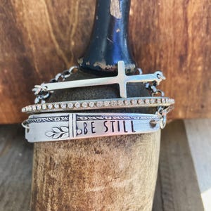 May include: A stack of silver bracelets with religious symbols. The top bracelet has a cross, the middle one has small clear stones, and the bottom one says "BE STILL" with a leaf design. The bracelets are displayed on a wooden surface.
