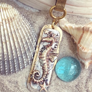 May include: A seahorse pendant necklace with a brass-colored seahorse design, set against a silver background. The pendant is rectangular with a gold-colored border and hangs from a ball chain. A blue glass bead and seashells are in the background.