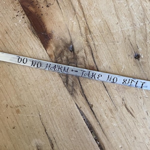 Op de afbeelding: Een zilverkleurige metalen armband met de woorden "DO NO HARM" en "TAKE NO SHIT" erin gestempeld. Een pijldesign scheidt de twee zinnen. De armband wordt gepresenteerd op een houten oppervlak.