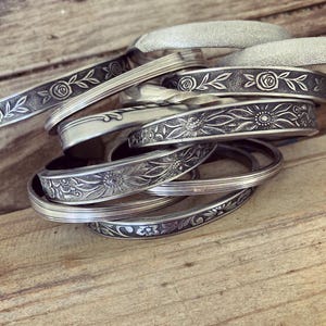 May include: A collection of silver-toned metal bangles featuring various embossed patterns including floral motifs, roses, and sunflowers. Some bracelets have a smooth polished finish, while others display a textured or twisted surface design.