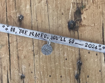 May include: A silver metal bracelet with the text "Oh, the places you'll go - 2024" engraved on it. A small silver compass charm hangs from the bracelet.