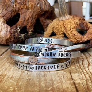 Op de afbeelding: Een set van vijf zilveren metalen armbanden met Halloween-gravures. De armbanden bevatten de zinnen "Boo", "Bump in the Night", "Hocus Pocus" en "Happy Halloween" samen met afbeeldingen van geesten, vleermuizen en pompoenen.