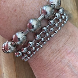 May include: A set of four silver bracelets with different sized ball chain links. The bracelets are stacked on a person's wrist.