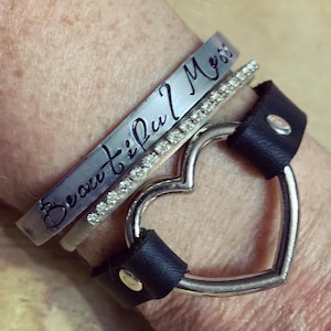 May include: A set of three silver bracelets, one with the inscription "Beautiful Mess", one with a row of clear rhinestones, and one with a heart-shaped design. The bracelets are worn on a person's wrist.