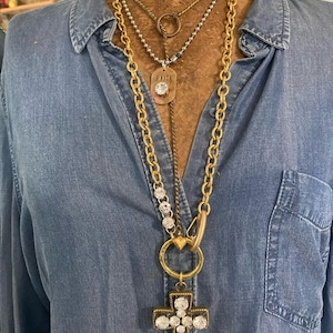 May include: A collection of layered necklaces with gold and silver chains. The focal point is a large gold chain with a cross pendant encrusted with clear crystals. Other necklaces include a pendant with the word "Faith".