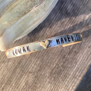 May include: A silver and gold cuff bracelet with the names "Rowan" and "Maverick" engraved on it, separated by a small gold heart charm.