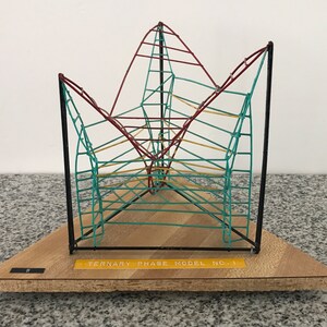 May include: A colorful, geometric model labeled "TERNARY PHASE MODEL NO. 1" on a wooden base. The model features a wireframe structure with red, teal, and yellow lines, supported by black vertical posts. The base is rectangular and made of light-colored wood.