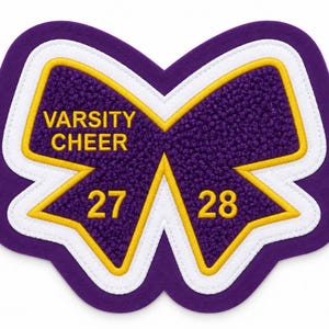 May include: A purple and white chenille patch shaped like a cheer bow with yellow embroidery. The text VARSITY CHEER is on the left loop, with the numbers 27 and 28 on the bottom tails. The patch features a thick purple border and yellow trim.
