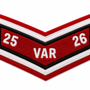 May include: A chevron-shaped embroidered patch featuring a red background with a white and black border. The patch displays the white embroidered text 25 and 26 on the upper sides and the white text VAR centered at the bottom point of the chevron.
