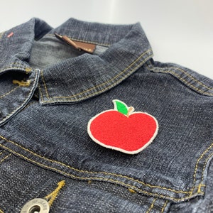 Puede incluir: Un parche bordado de una manzana roja con un tallo y una hoja verdes sobre una chaqueta vaquera azul oscuro.