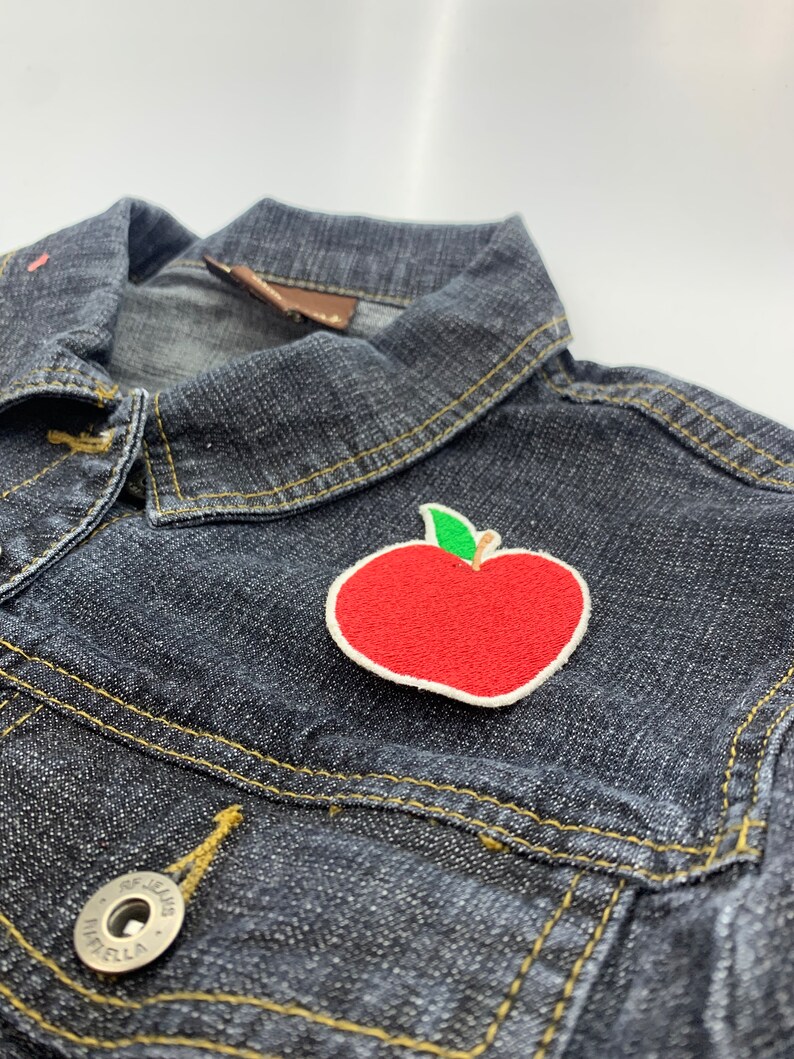 Puede incluir: Un parche bordado de una manzana roja con un tallo y una hoja verdes sobre una chaqueta vaquera azul oscuro.