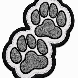 May include: An embroidered patch featuring two overlapping animal paw prints. The design consists of textured grey pads and toes outlined in white, with a thick black border surrounding the entire shape. The stitching is dense and uniform throughout.