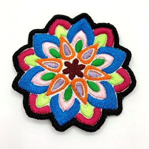 May include: A colorful embroidered patch with a flower design in shades of pink, blue, green, orange, and brown. The patch is surrounded by a black border.