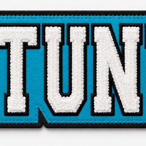 May include: A rectangular embroidered patch featuring the word STUNT in bold white block letters with a black outline. The background of the patch is bright cyan blue, and the entire piece is finished with a thick black stitched border.