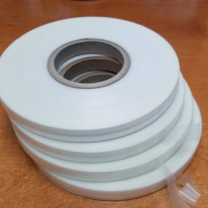 May include: A stack of white adhesive tape rolls, with the tape unwinding from the bottom roll.