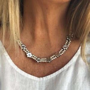 May include: A silver necklace with a floral design. The necklace features small, silver flower-shaped beads and round beads, creating a layered effect. The necklace is worn over a white, V-neck top.