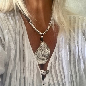 Könnte beinhalten: Halskette mit einem silberfarbenen Anhänger in Form einer strukturierten Austernschale. Die Kette hat eine dunkelbraune Kordel und silberne Perlen. Die Person trägt ein weißes Hemd mit Knöpfen.