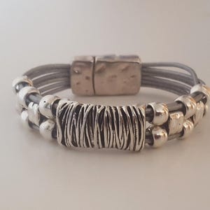 Puede incluir: Un brazalete de cuero gris con cuentas plateadas y un cierre de metal plateado. El brazalete tiene un diseño tejido y está hecho de varias hebras de cuero.