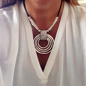 Könnte beinhalten: Eine silberne Halskette mit einem großen, runden Anhänger aus mehreren konzentrischen Ringen. Der Anhänger hat eine strukturierte Oberfläche und ist an einem schwarzen Lederband befestigt.